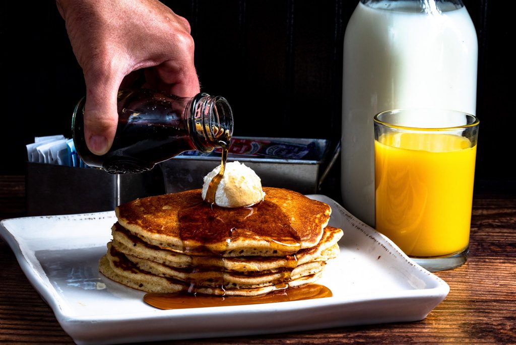 Stack of pancakes on a white plate with a dollop of butter while syrup is poured from a small glass bottle, next to a glass of orange juice and a bottle of milk.