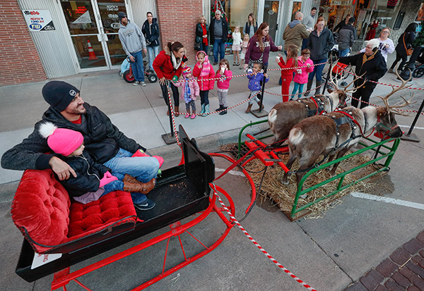a father and daughter sitting on a sled with two reindeer as other families wait in line.