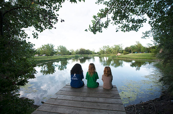 three girls sitting on edge of wood dock facing the lake. 