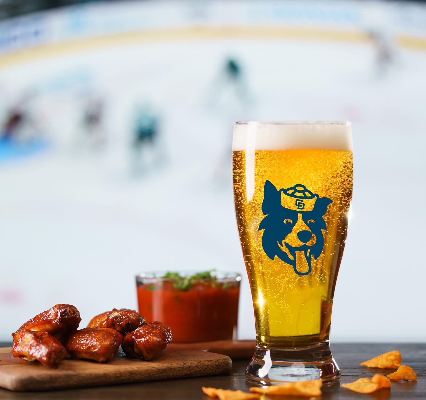 Glass of beer with a blue logo showing a dog’s face on it on a table next to a wooden board of sauced chicken wings, a small bowl of red sauce, and scattered chips.