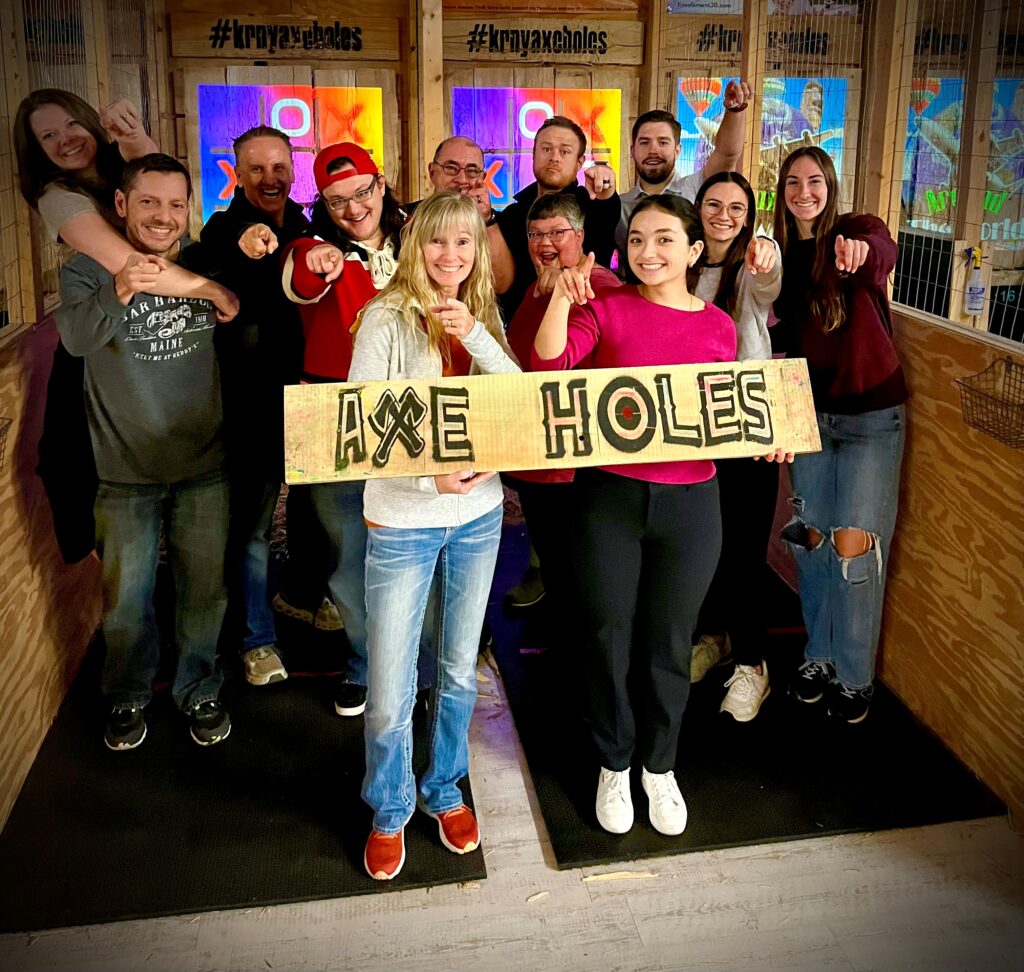 People holding Axe Holes wood sign while pointing towards the camera. 