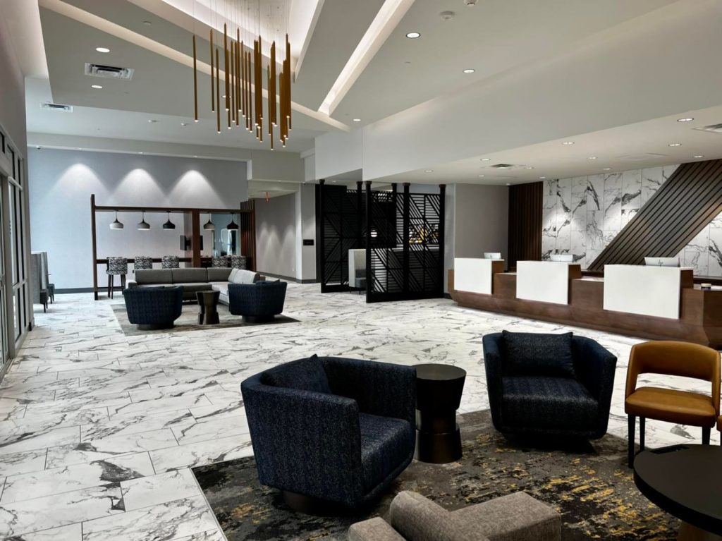Modern lobby with marble flooring, black cushion chairs, hanging lights and reception desk alongside wall.