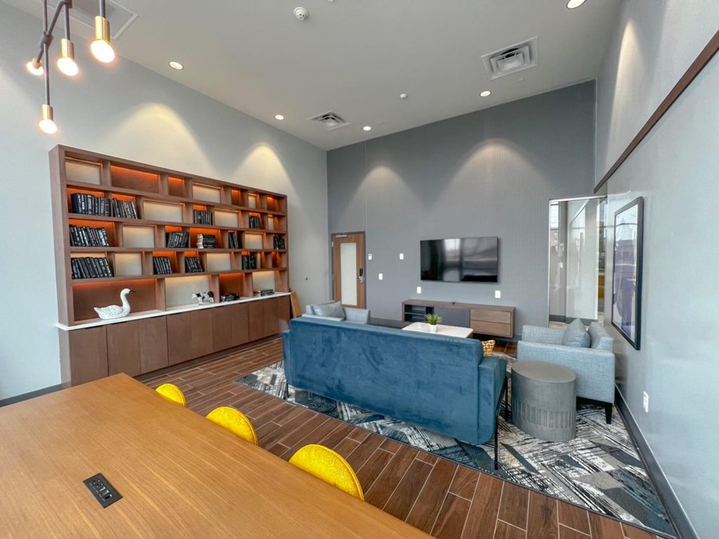 grey room with blue couch facing tv, a table with yellow chairs, and wall with shelves of books and decor.