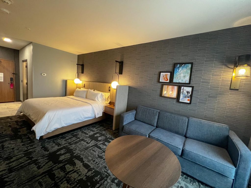 hotel room with white bedspread, handing lights at bedside with a blue couch and wood table and artwork on walls.