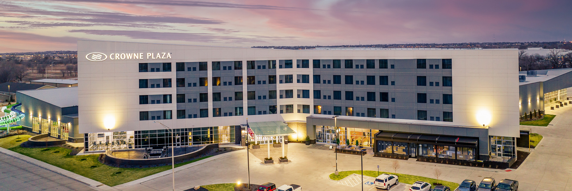 Crowne Plaza hotel building with its “CROWNE PLAZA” sign on the facade, multiple floors of windows, vehicles parked in front, and adjacent structures under a cloudy sky.