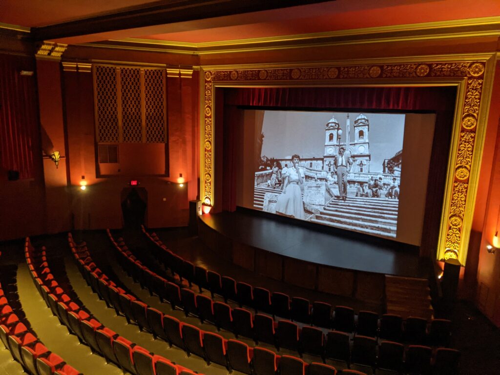Historic interior of the World Theater with red walls and cushioned seating, gold decorated archway and a black and white movie playing on the screen.