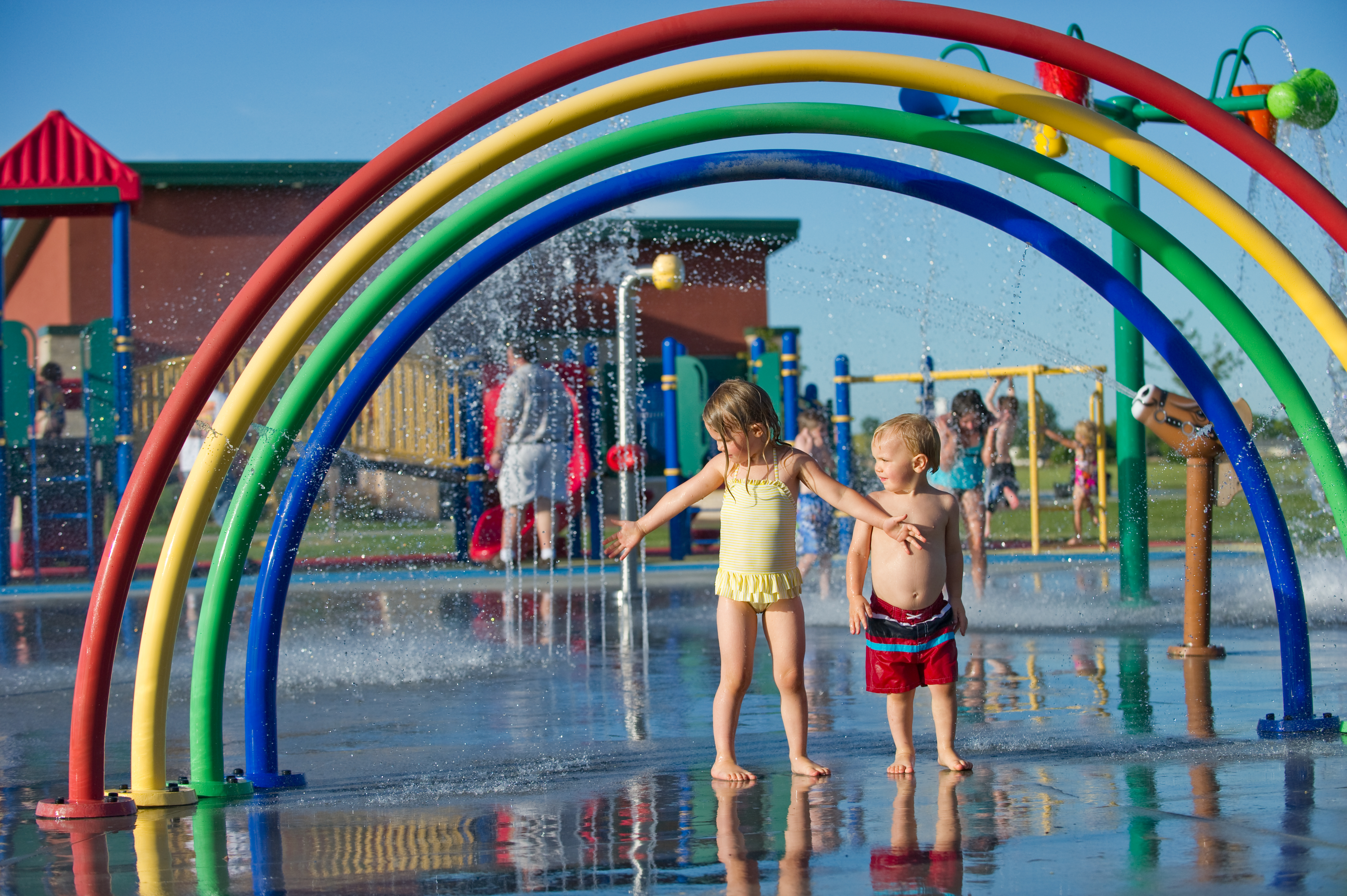 Children in swimsuits standing under curved, rainbow-colored water arches at an outdoor Kearney splash pad at Yanney Park