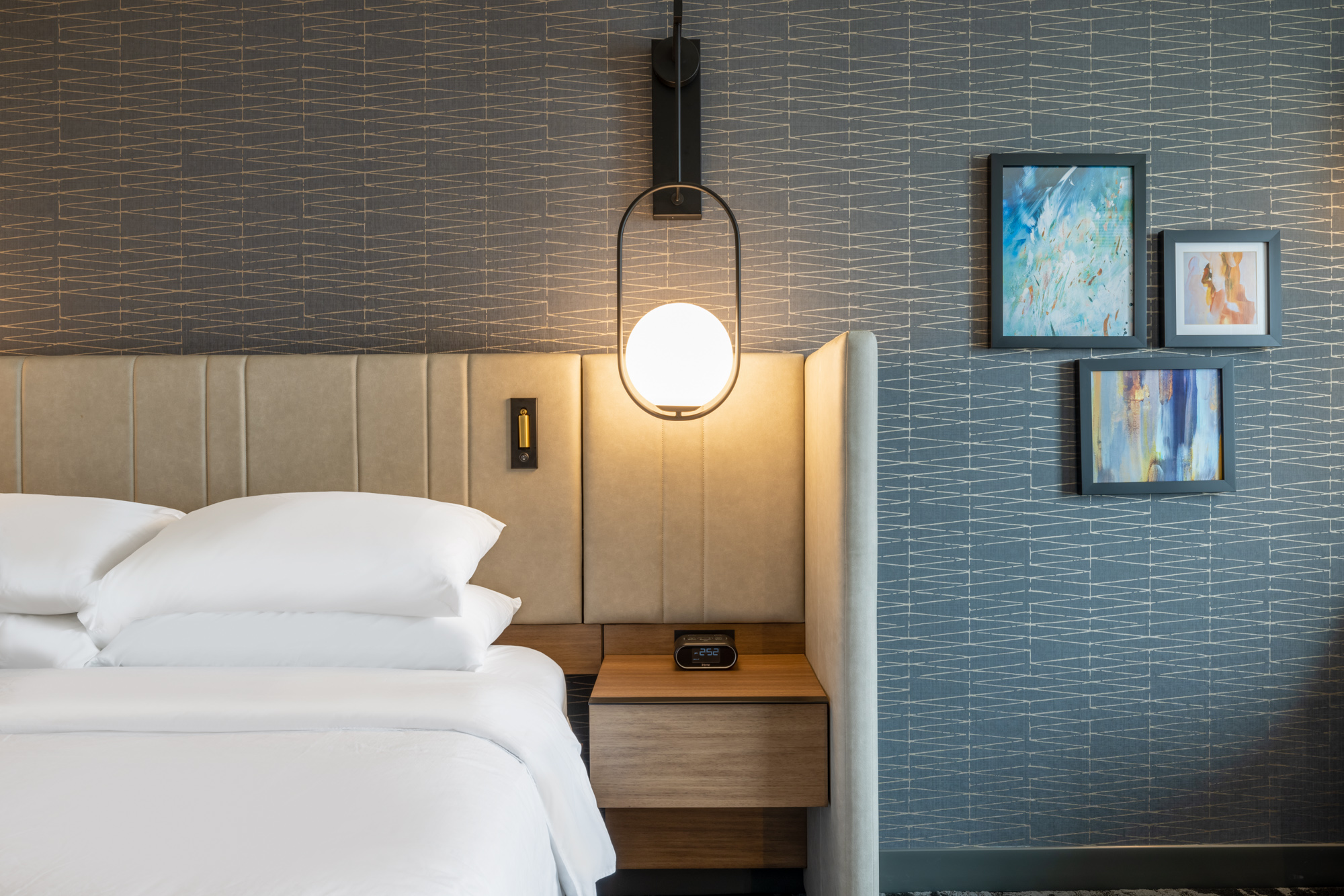 Queen-sized bed with white linens, a wooden nightstand holding a hanging bedside lamp, and abstract framed wall art visible behind the bed.