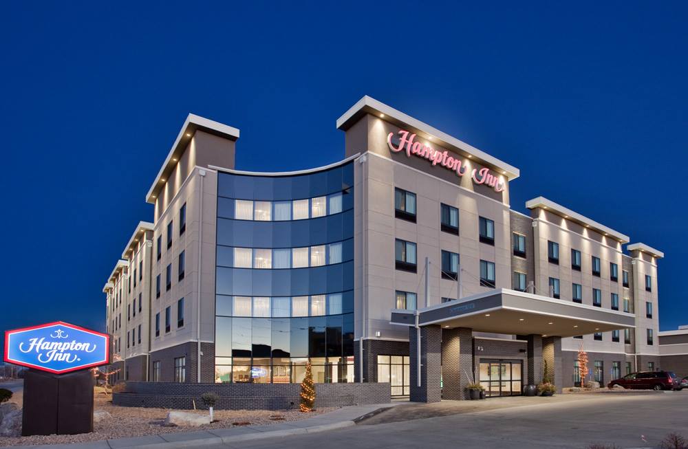 Neutral exterior view of a multi-story Hampton Inn hotel building with rows of windows, a main entrance canopy, and a parking area in front under a clear sky.