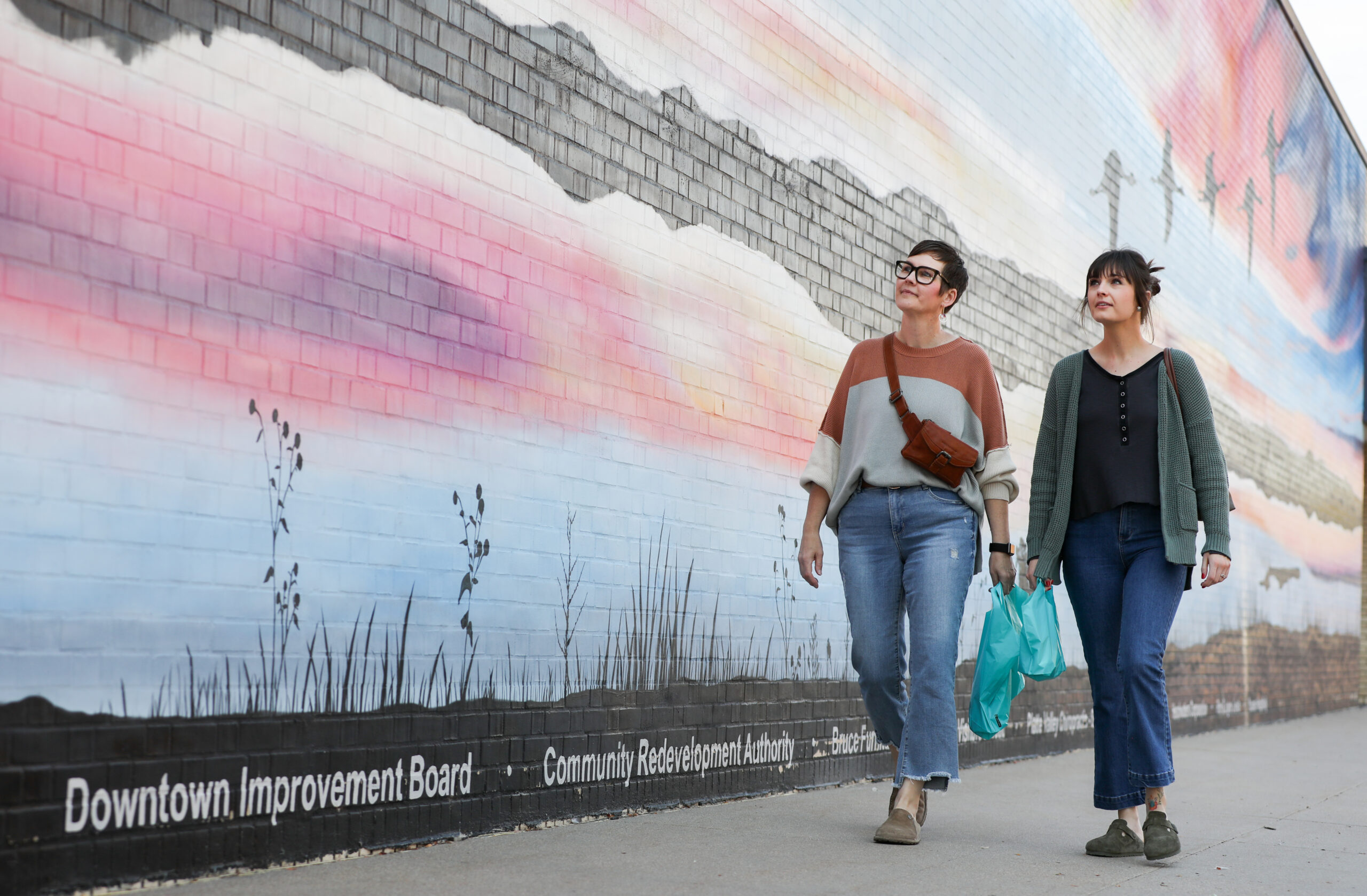 Two women walking down sidewalk along mural brick wall.