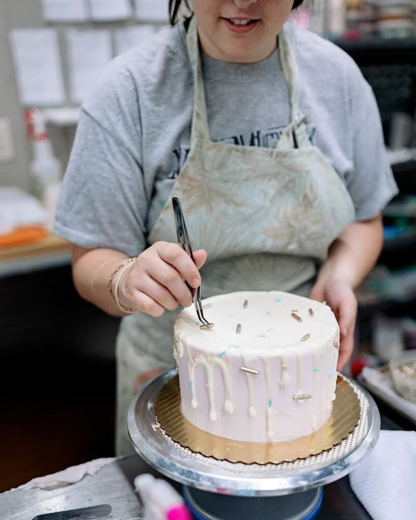 Person wearing apron as they places sprinkles on decorated cake. 