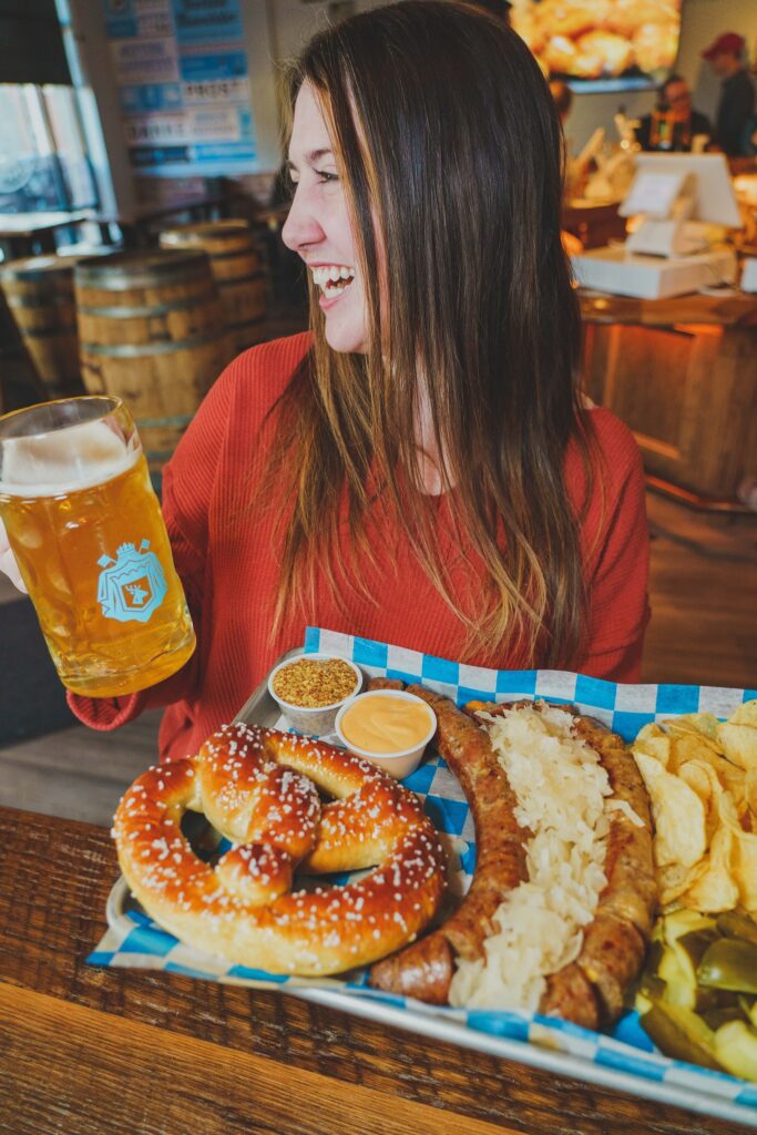 person smiling while holding beer mug and tray loaded with a pretzel, chips, pickles, bratwursts and sauerkraut. 