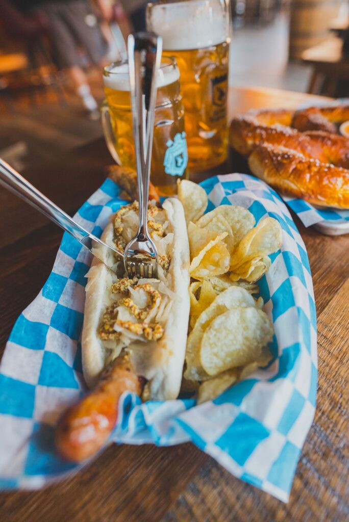 loaded bratwurst with side of chip and nacho cheese. Two glass mugs of beer on table.  
