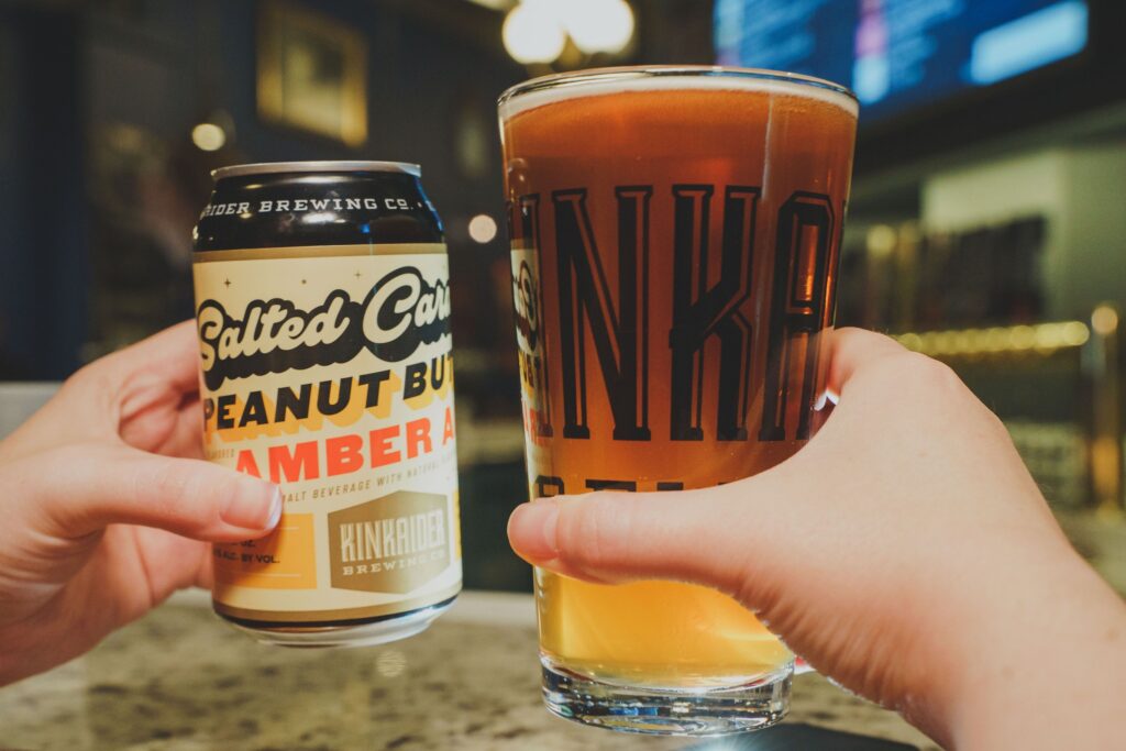 Two handing holding a glass of salad caramel and peanut butter flavored beer and a glass of beer. 