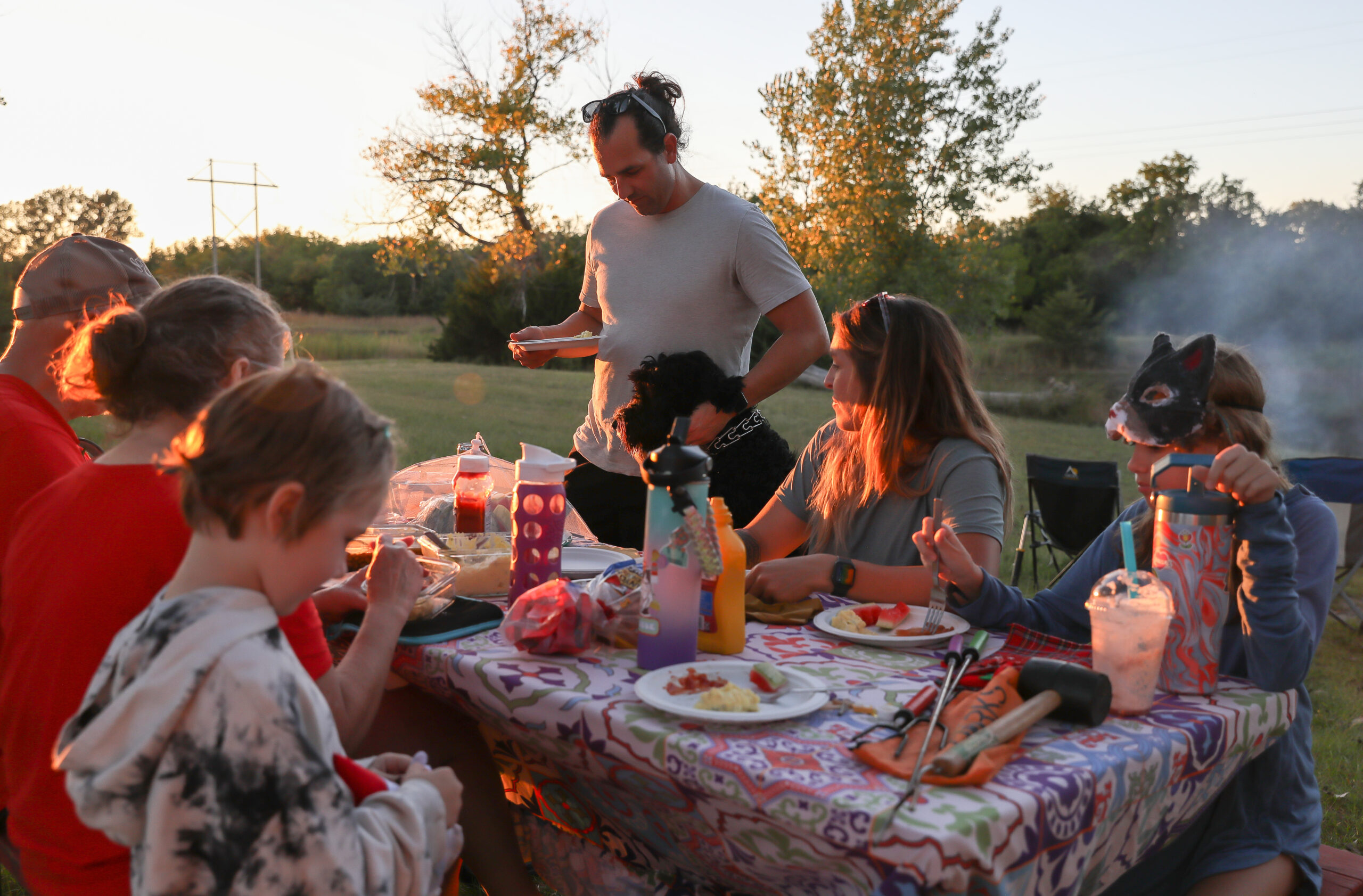 A family sitting on a picnic table with plates of food as they use an outdoor grill in the back.