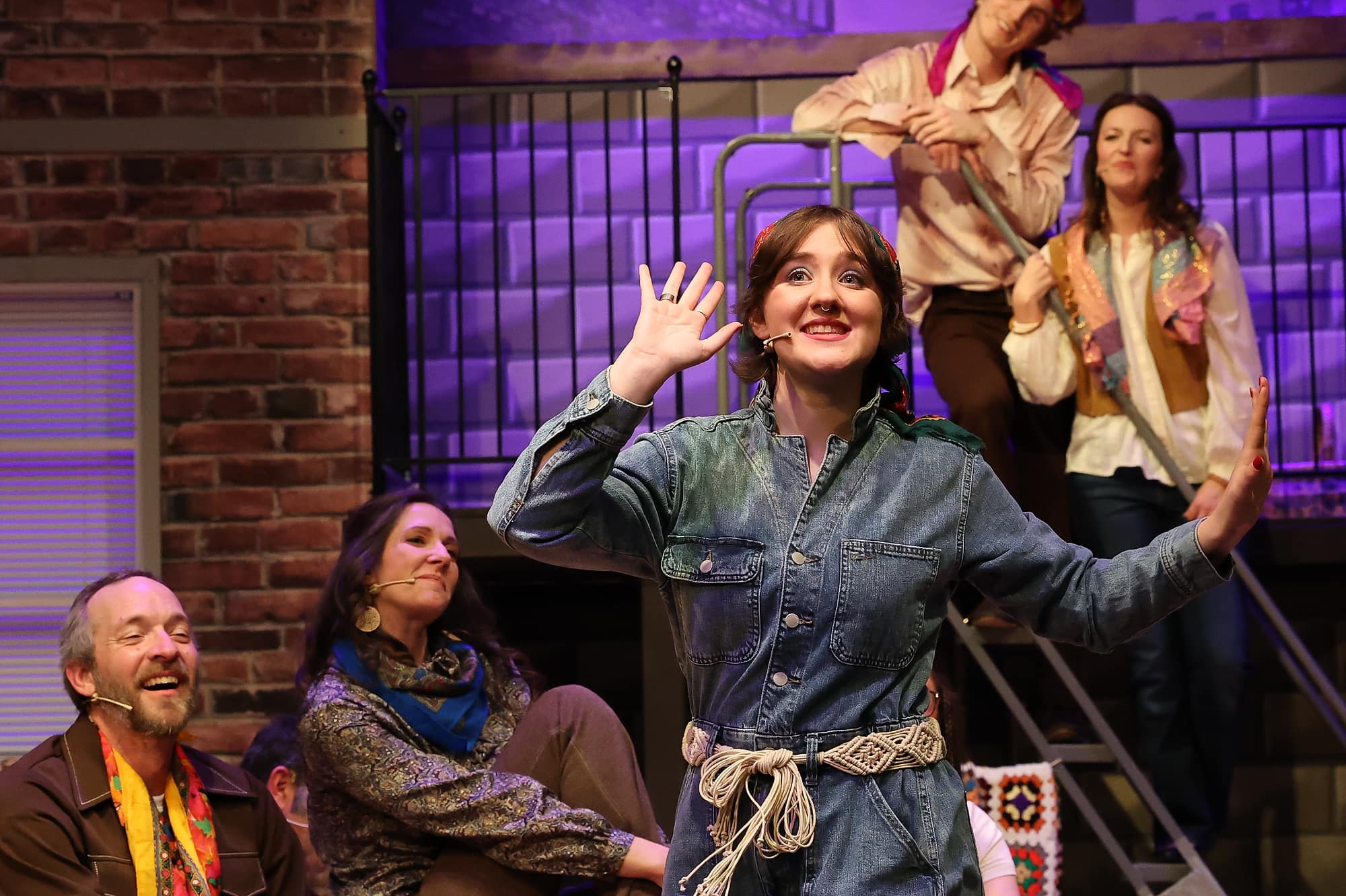Young performers on a theatre stage, with a person in a denim‑style outfit at center raising one hand while others sit and stand behind them in costume.