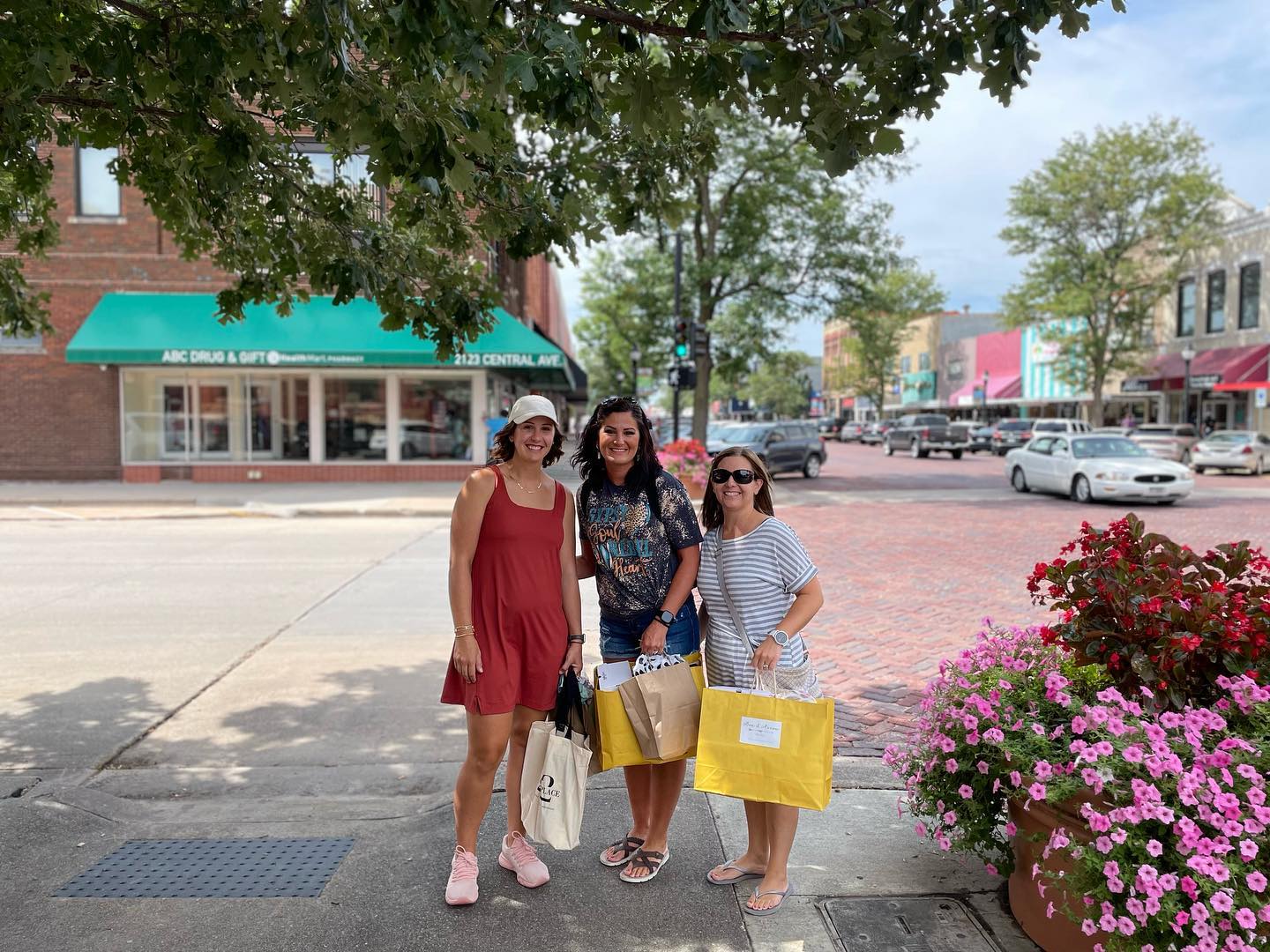 Three women standing on a sidewalk holding shopping bags with pink flowers and store fronts visible along the street.